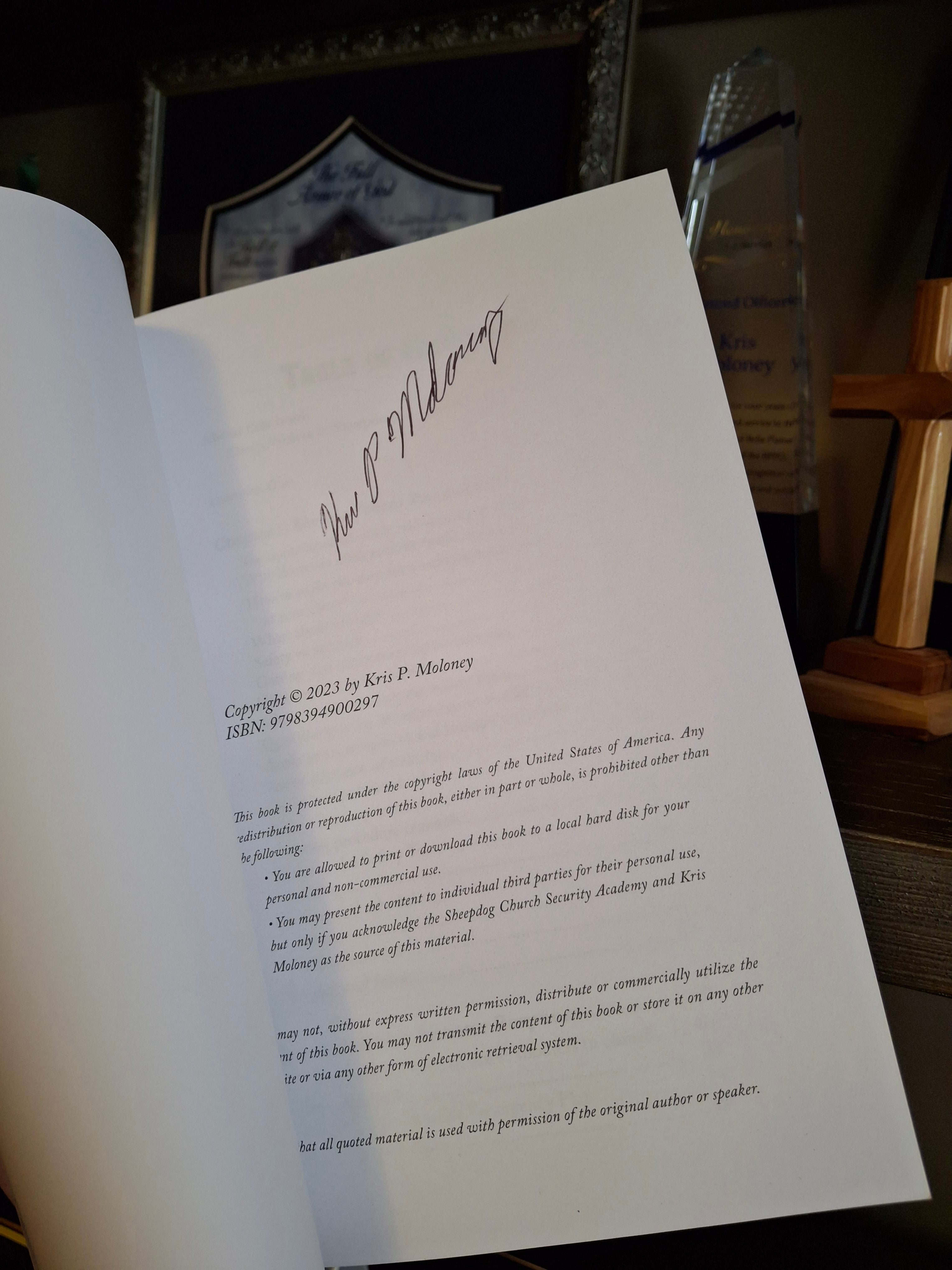 Inside page of Shepherding the Sheepdogs paperback showing Kris Moloney’s author signature on a signed copy, with wooden cross in background